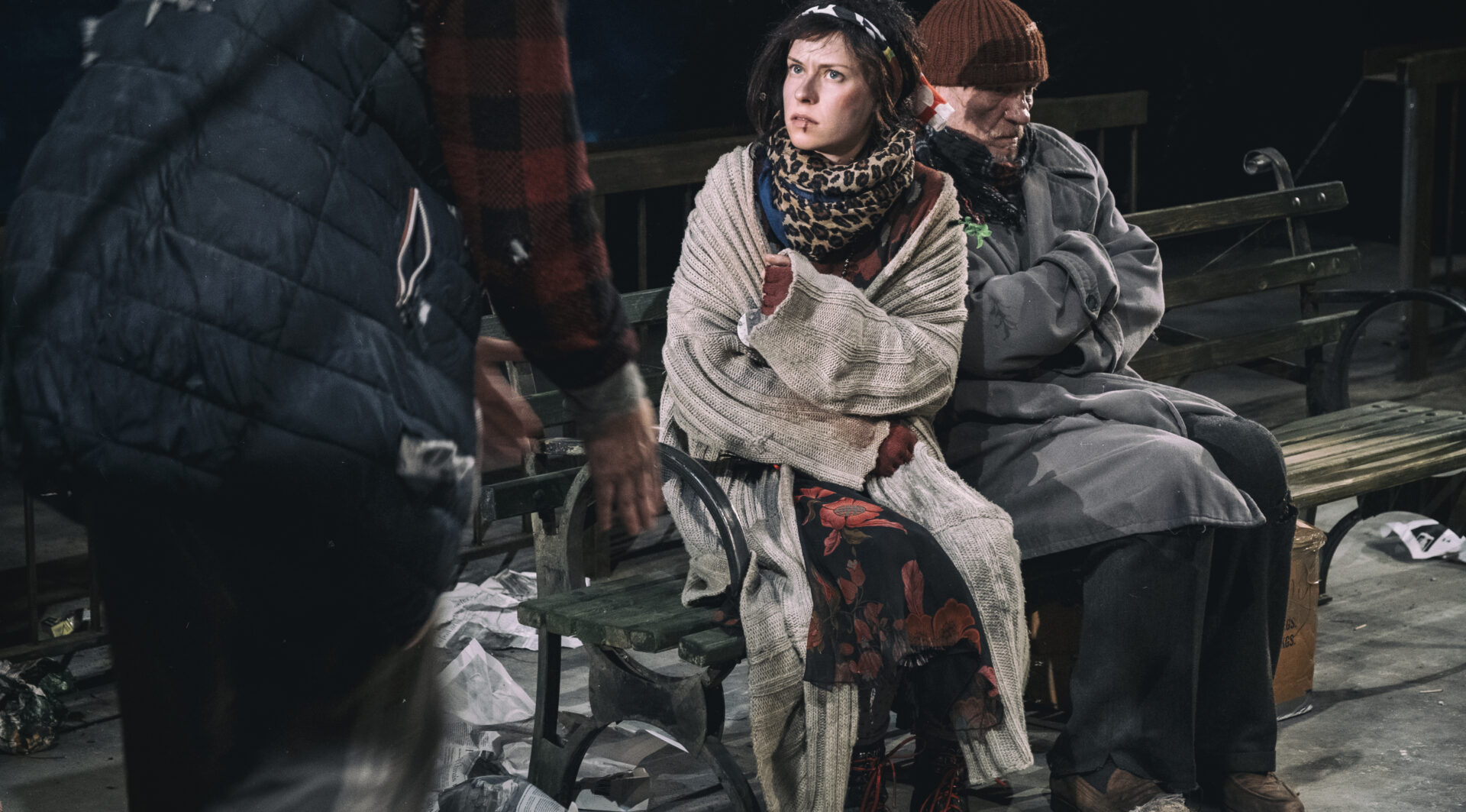 Są trzy osoby. Dwie siedzą na ławce. Kobieta i mężczyzna. Przed nimi stoi mężczyzna. Jest on zwrócony przodem do osób na ławce. Po lewej stronie ławki siedzi kobieta. Zwrócona jest w stronę mężczyzny. Prawą ręką wskazuje w jego stronę. Obok niej siedzi mężczyzna. Ma na sobie szary płaszcz. Mężczyzna patrzy przed siebie. Ręce ma założone na krzyż na klatce piersiowej.
