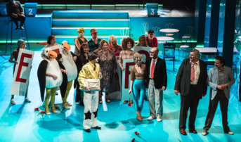 Kliknij by powiększyć zdjęcie. Alternatywny opis zdjęcia: Scena zbiorowa. Na scenie jest 19 aktorek i aktorów. Patrzą w różnych kierunkach. Wiekszość ubrana jest w kostiumy stylizowane na lata 80, ale 2 aktorki są w stroju pingwina. Ich uwaga skupiona jest na mężczyźnie w żółtej koszuli, który stoi z torbą na środku sceny. Scenografia jest niebieska. Za aktorami i aktorkami stoją stoliki z krzesłami. W tle schody.
