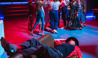 Kliknij by powiększyć zdjęcie. Alternatywny opis zdjęcia: Na skórzanej kanapie leży mężczyzna. Ma założone nogi na oparcie. Jedną reką trzyma kapelusz, a druga zwisa pozą kanapą. Za nim na scenie kilkanaście aktorek. Tworzą jedną grupę w okręgu.