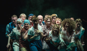 Kliknij by powiększyć zdjęcie. Alternatywny opis zdjęcia: Scena zbiorowa. W zwartej grupie stoi kilkanaście aktorek i aktorów. Mają poważne miny. Wszyscy patrzą się w przód. Śpiewają. Są ubrani w kostiumy stylizowane na lata 80.