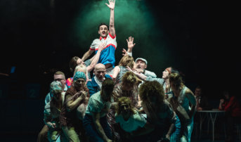 Kliknij by powiększyć zdjęcie. Alternatywny opis zdjęcia: Scena zbiorowa. 15 aktorów i aktorek tworzą zwartą grupę na środku sceny. Z tyłu najbardziej widoczny jest mężczynzna w koszulce polo. Jest wyżej niż wszyscy. Ma wyciągnięta lewą rękę ku górze.