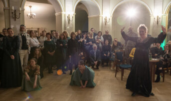 Kliknij by powiększyć zdjęcie. Alternatywny opis zdjęcia: Scena na Foyer I piętra w teatrze. W półkolu stoi widownia. Na pierwszym planie kobieta w zielonej sukni z uniesionymi rękami, wyglądająca na prowadzącą wydarzenie. Na podłodze przed nią dwie młode kobiety w zielonych strojach kucają. W tle eleganckie łuki i kinkiety na ścianach.