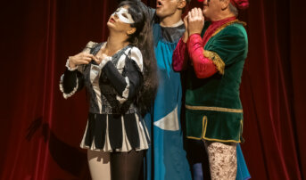 Kliknij by powiększyć zdjęcie. Alternatywny opis zdjęcia: Trójka aktorów w maskach i kolorowych strojach teatralnych pozuje w dynamicznej pozie na tle czerwonej kurtyny.