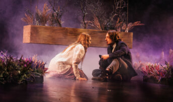 Kliknij by powiększyć zdjęcie. Alternatywny opis zdjęcia: Młoda kobieta [Mary] i młody chłopak [Dick] siedzą pomiędzy wrzosowiskiem. Za nimi zwisa z sufitu podłużna skrzynia. Jest w niej mini ogród. Kobieta ubrana jest w jasną sukienkę, która na dole jest fioletowa. Ma na szyi klucz. Chłopak patrzy na nią. Jest ubrany w ciemny kostium.