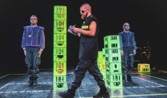 Na scenie otoczonej neonowymi liniami znajduje się kilka wież z plastikowych skrzynek. Mężczyzna w czerni z brodą przechodzi przed dwójką aktorów w uniformach.