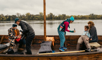 W drewnianej łodzi pięć osób. Mężczyzna w kolorowej kurtce siedzi na ławce z plecakiem. Kobieta w niebieskiej kurtce trzyma kamerę i robi zdjęcie mężczyźnie w jasnej koszuli, który siedzi z opartą głową. Inna osoba trzyma czerwoną torbę i pomaga w organizacji sprzętu.