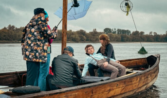 Wnętrze łodzi z pięcioma osobami. Kobieta z niebieskim parasolem trzyma kamerę, podczas gdy mężczyzna w ciemnej kurtce rozkłada sprzęt fotograficzny. Dwie inne osoby, jedna w czarnej piance, a druga w jasnej koszuli, przygotowują się do zdjęcia. W tle jest spokojna woda.