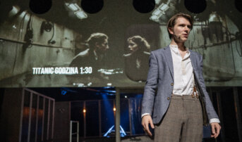 Kliknij by powiększyć zdjęcie. Alternatywny opis zdjęcia: Mężczyzna w szarym garniturze na tle projekcji z napisem „TITANIC GODZINA 1:30”. Na ekranie jest projekcja wygenerowana przez sztuczną inteligencję na której jest para w podtapianym statku.