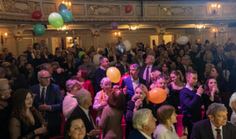 Widownia. Wszyscy trzymają w dłoniach szampana. Z góry spadają kolorowe balony.