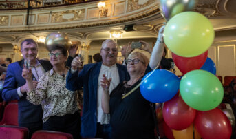 Dwie pary stoją na widowni i trzymają w dłoniach szampana. Kobieta po prawej stronie trzyma związane balony.