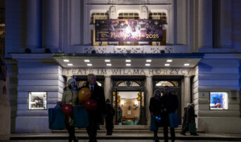 Goście wychodzą a budynku teatru. Trzymają w dłoniach upominki. Na fasadzie teatru grafika z życzeniami noworocznymi.