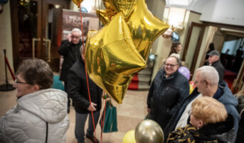 Goście tłoczą się w przedsionku teatru. Trzymają w dłoniach upominki. Na wejściu wiszą balony.