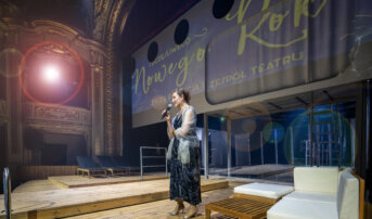 Na scenie stoi kobieta (Renata Derejczyk - dyrektorka teatru) z mikrofonem. Nad nią na ekranie wyświetla się grafika z życzeniami noworocznymi.