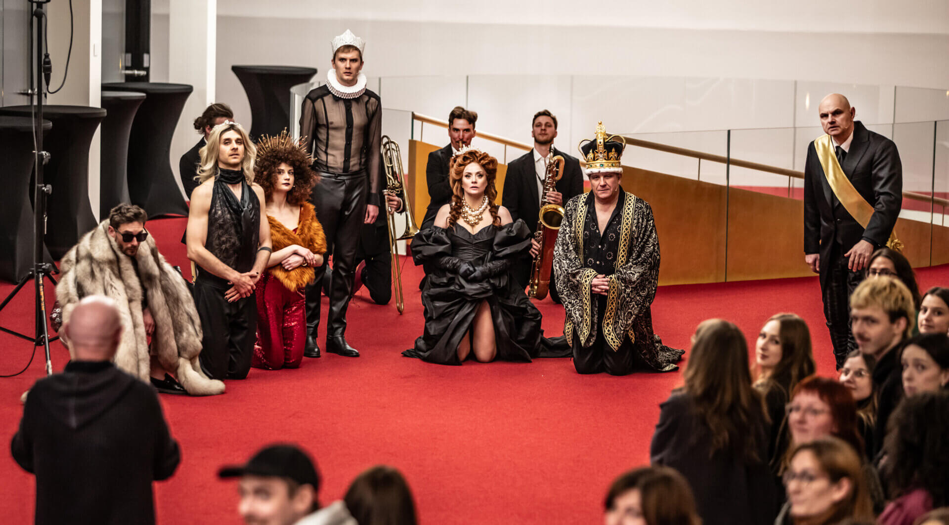Kliknij by powiększyć zdjęcie. Alternatywny opis zdjęcia: Grupa aktorów na czerwonym dywanie. Kobieta w czarnej sukni i mężczyzna w koronie klęczą na pierwszym planie, za nimi muzycy i postacie w ekstrawaganckich kostiumach.