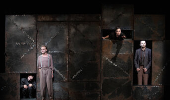 Kliknij by powiększyć zdjęcie. Alternatywny opis zdjęcia: Cztery osoby znajdują się w różnych otworach dużej, zardzewiałej konstrukcji. Na dole stoi boso mężczyzna w luźnych spodniach i szelkach. Po lewej stronie ktoś kuli się w małej przestrzeni. Na górze kobieta wyciąga rękę.