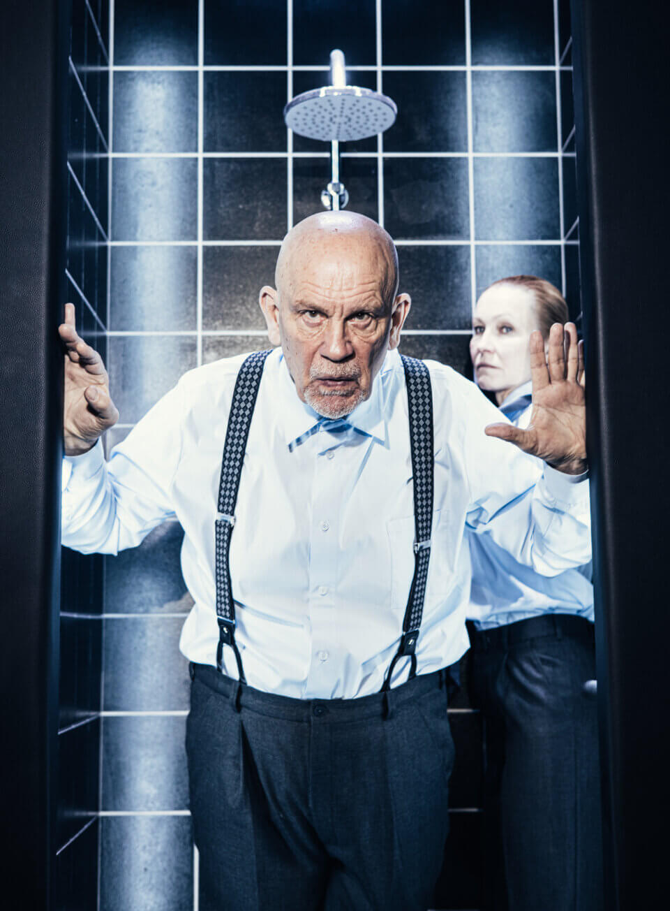 John Malkovich stoi w progu kabiny prysznicowej, dłonie na framudze. Za nim Ingeborga Dapkunaite. Czarne kafelki i deszczownica tworzą tło.