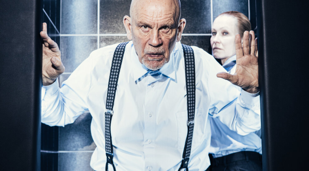 John Malkovich stoi w progu kabiny prysznicowej, dłonie na framudze. Za nim Ingeborga Dapkunaite. Czarne kafelki i deszczownica tworzą tło.