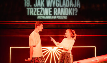 Kliknij by powiększyć zdjęcie. Alternatywny opis zdjęcia: Kobieta i mężczyzna stoją naprzeciw siebie na scenie z czerwonym światłem. Kobieta gestem palca zwraca się do niego. W tle wyświetlone jest pytanie: „Jak wyglądają trzeźwe randki?”