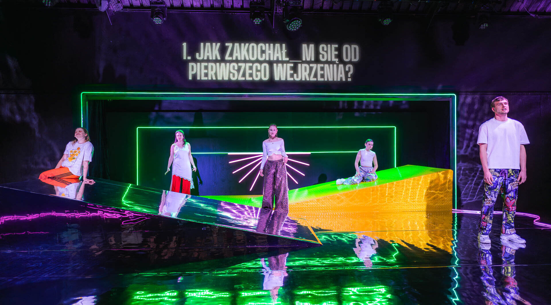 Kliknij by powiększyć zdjęcie. Alternatywny opis zdjęcia: * Pięcioro aktorów stoi na scenie z lśniącą podłogą i neonową scenografią. Nad sceną wyświetlone jest pytanie: „Jak zakochał_m się od pierwszego wejrzenia?”